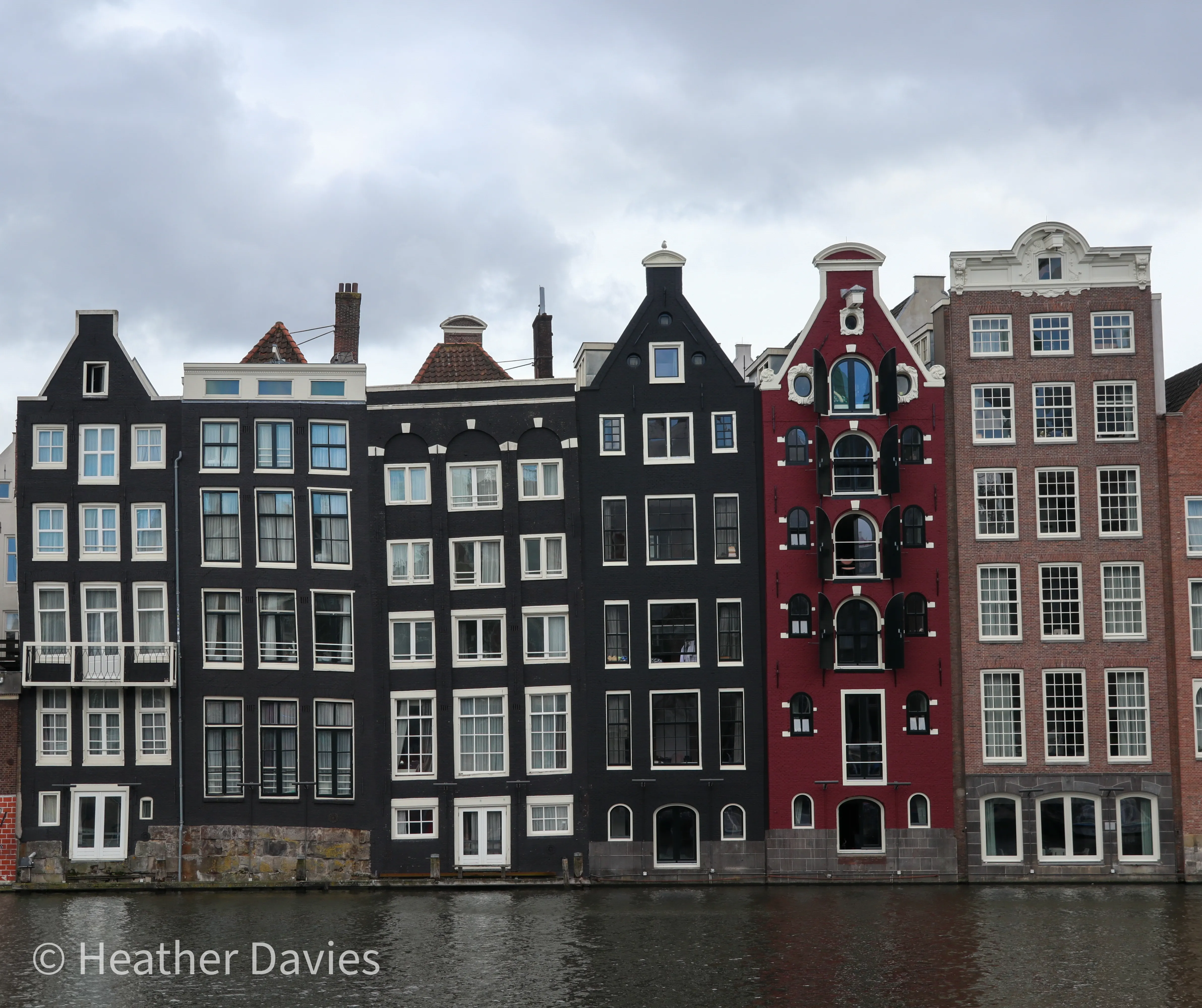 A row of narrow black, red and brick canal houses beneath a cloudy sky