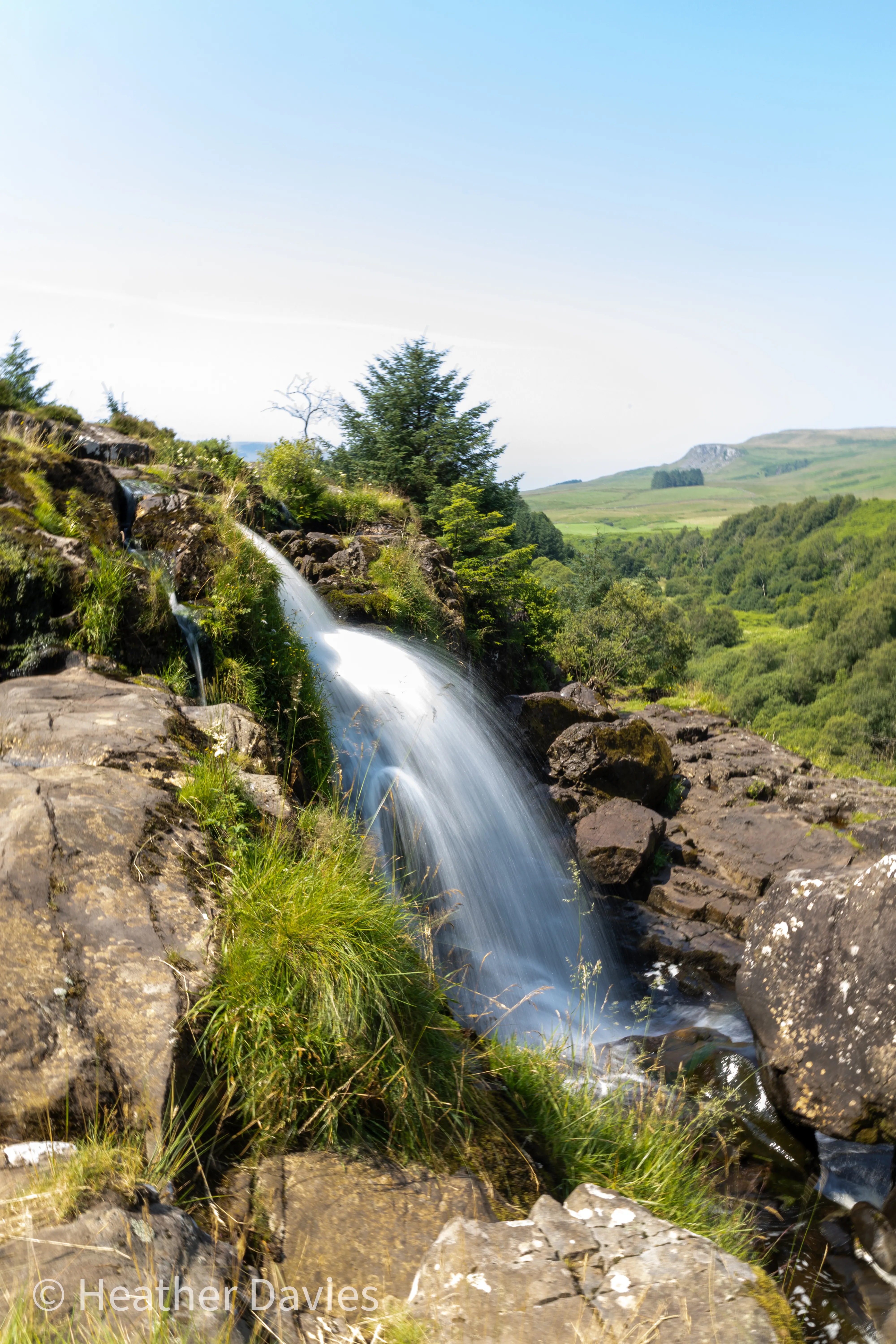 A waterfall flowing over rocks with the green Campsie hills beyond